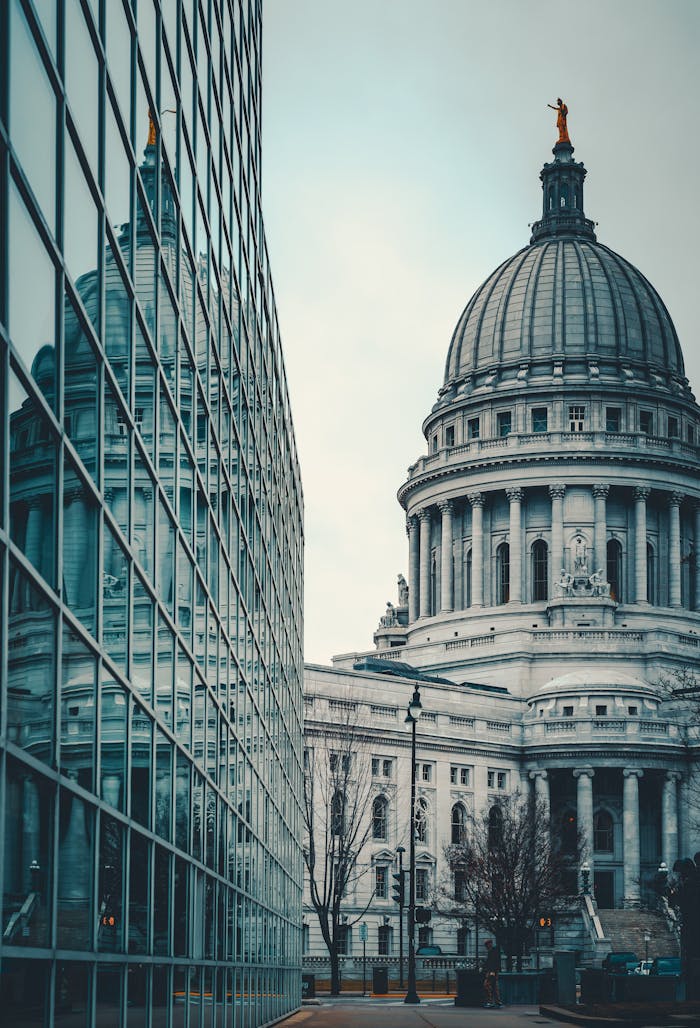 A modern glass building reflecting a historic capitol building in an urban setting, captured during the day.